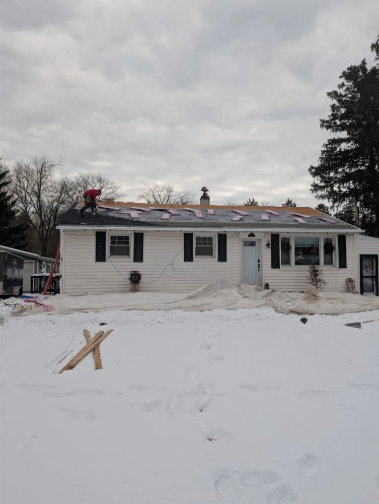 A roofer laying underlayment and shingles across a stripped roof in the snow